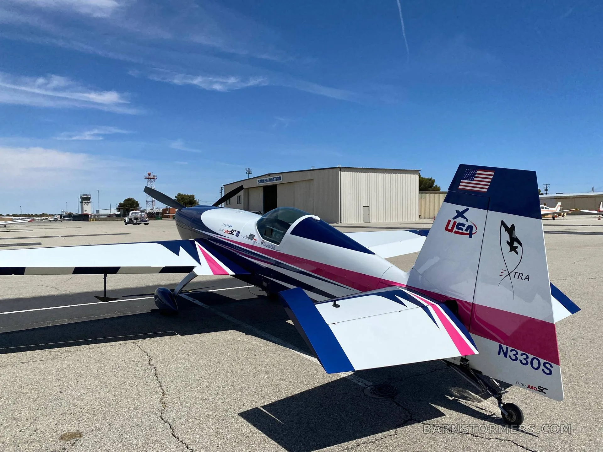 The rear view of an Extra 330SC aircraft with a pink, blue, and white paint scheme, parked on an airfield tarmac.