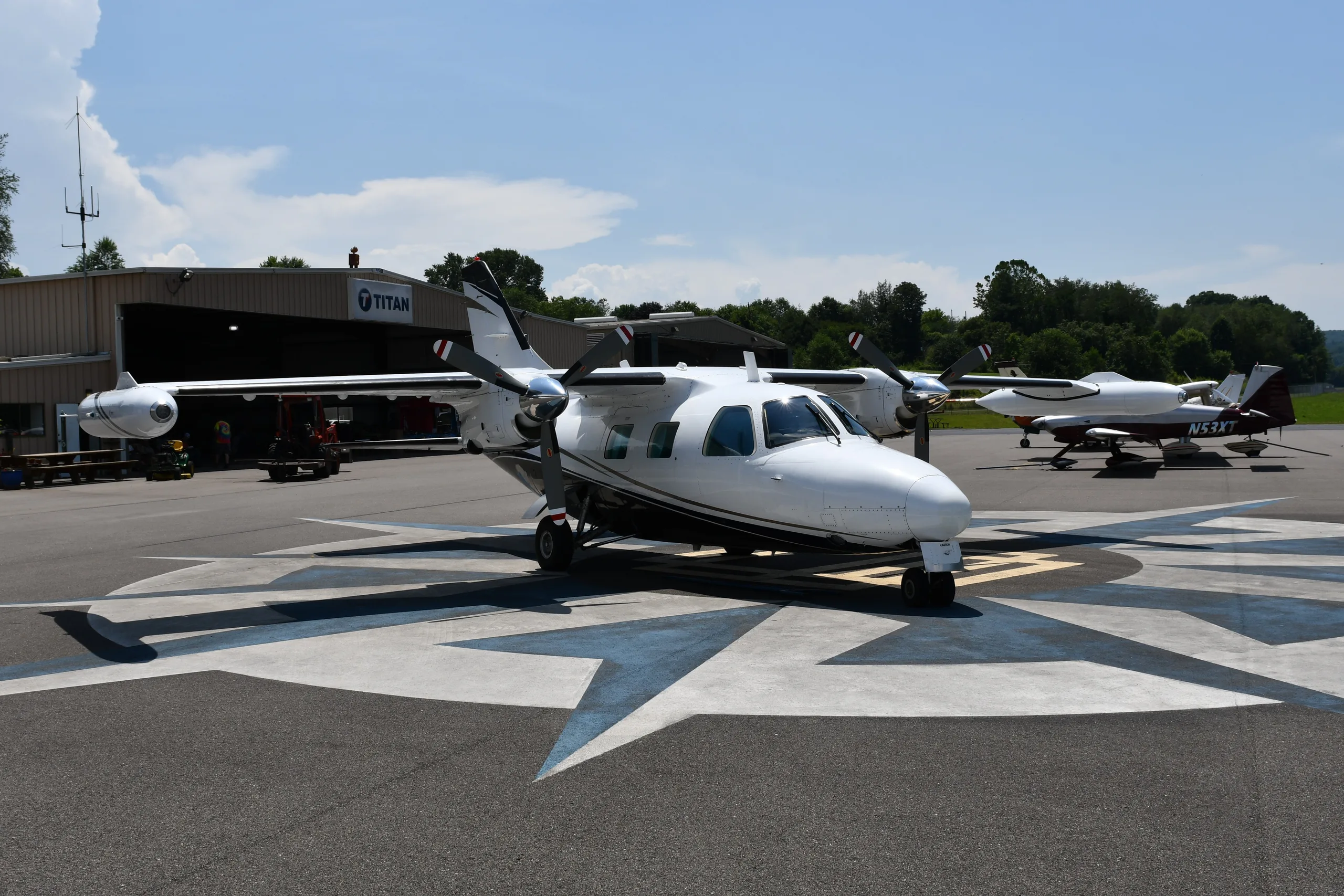 A white twin-engine propeller aircraft with dark stripes is parked on a compass rose at an airfield, with a hangar and trees in the background.