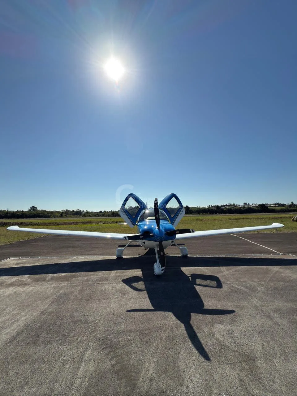 A front view of a blue and white Cirrus SR22 aircraft with its doors open, parked on an airfield under a bright sun.
