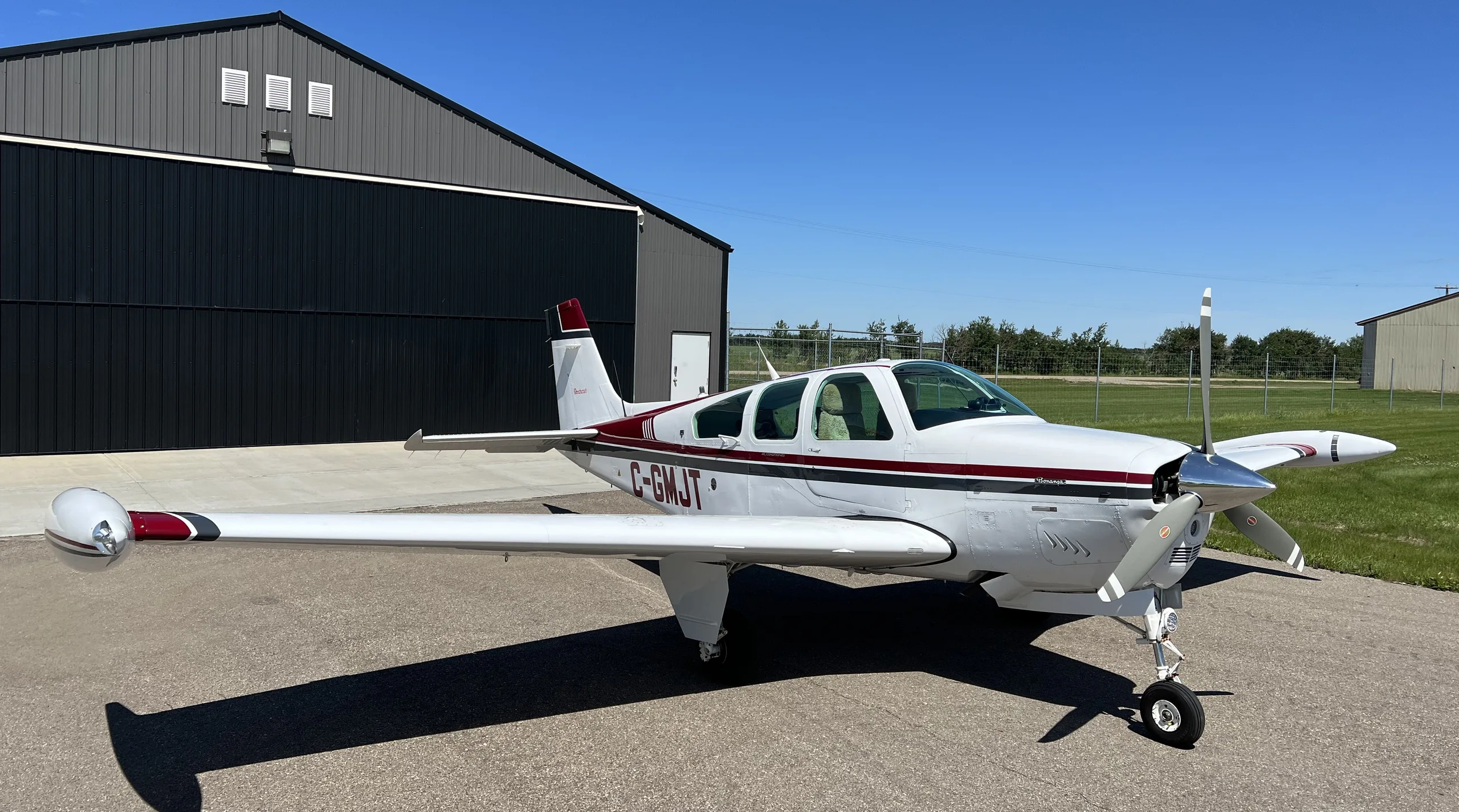 A white Beechcraft Bonanza with maroon and black stripes, registration C-GMJT, is parked on an airfield in front of a hangar under a clear blue sky.