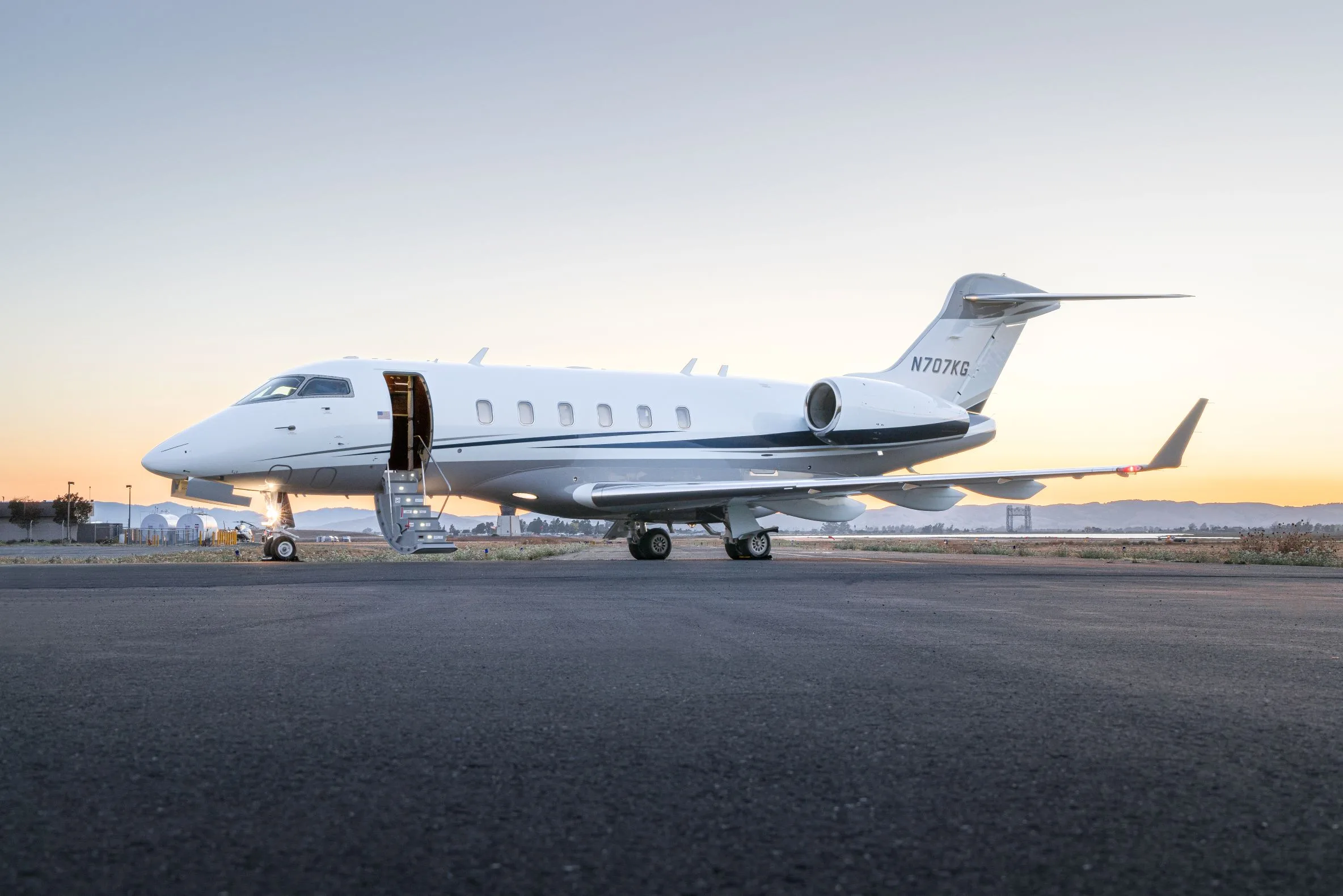 A white and grey business jet with its door open and stairs extended, parked on an tarmac at sunset.