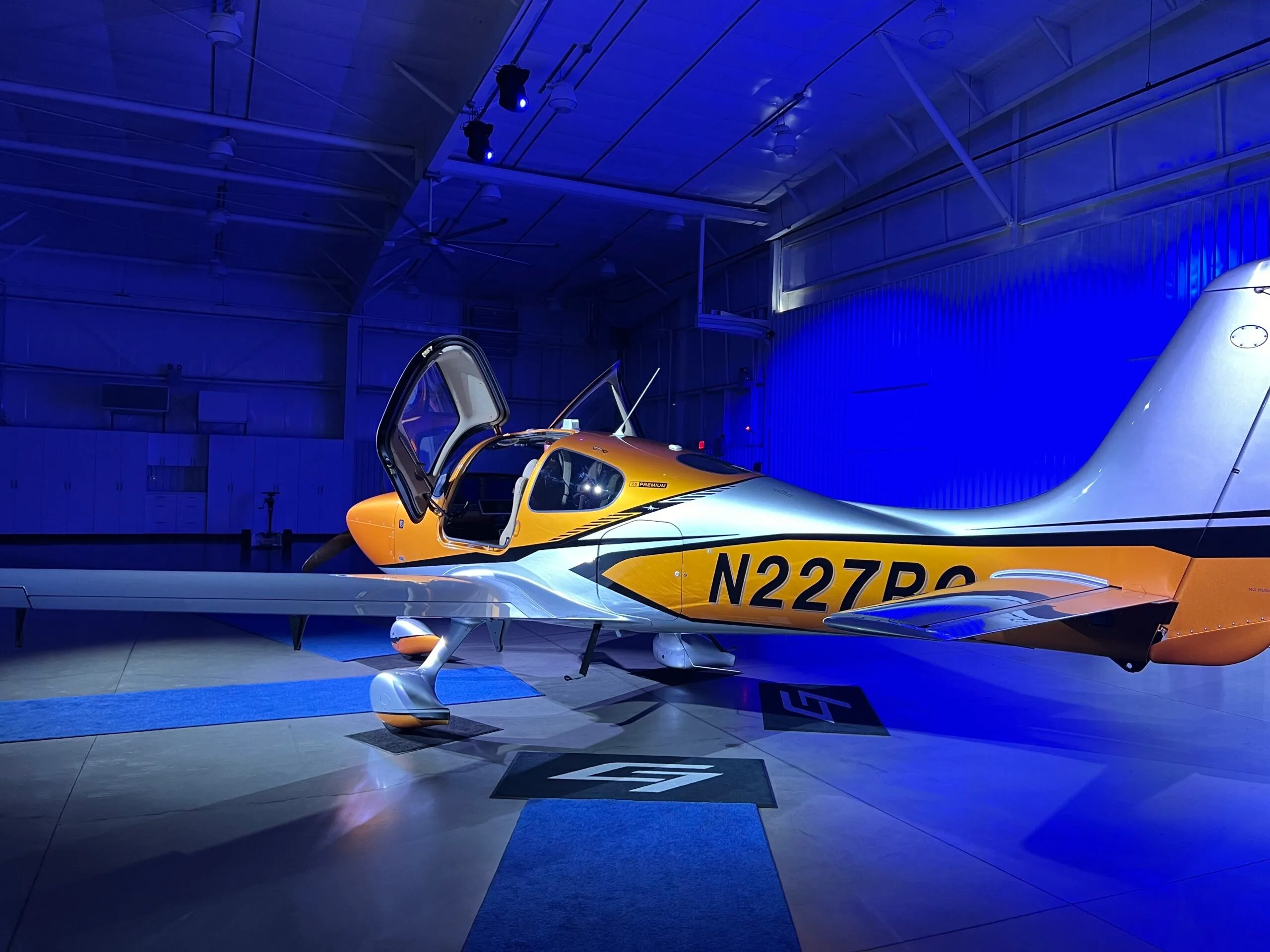 A Cirrus SR22 aircraft with orange and silver paint, doors open, on a blue carpet in a hangar with blue lighting.