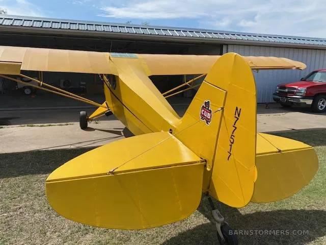 A rear view of the yellow Piper Cub-style aircraft, showing the tail with "N77517" and a "CUB" logo, parked in front of a hangar.