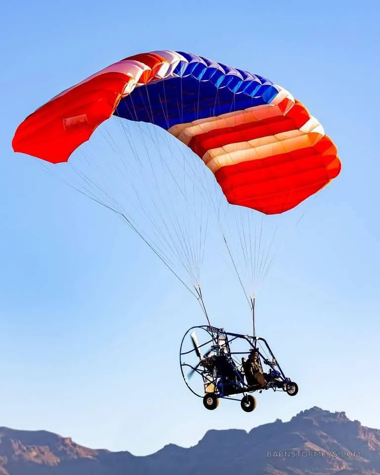 A powered parachute with a red, white, and blue canopy flies against a clear blue sky with mountains in the background.