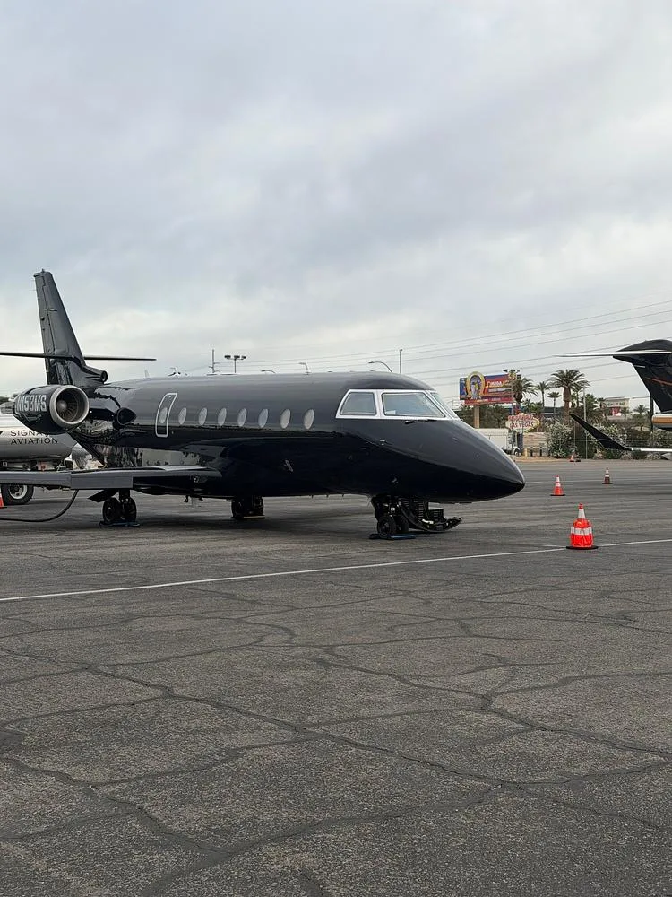 A black business jet, tail number N153MS, parked on an airfield with orange cones and distant buildings under an overcast sky.