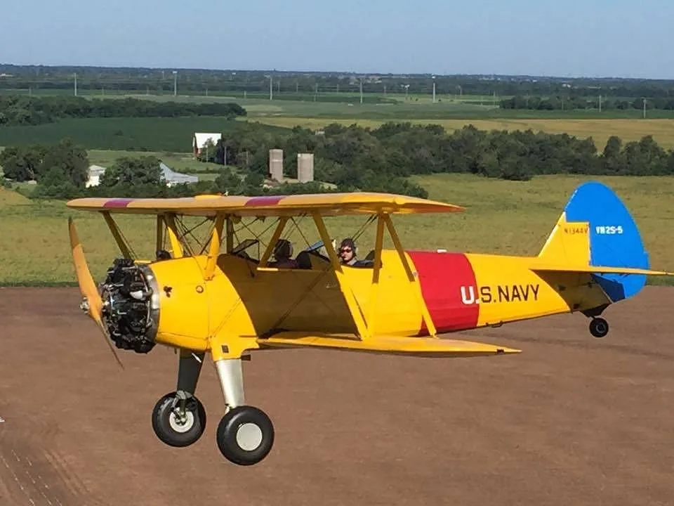 A yellow Stearman biplane with red and blue accents, marked 'U.S. NAVY' and N1344V, flying low over green fields.