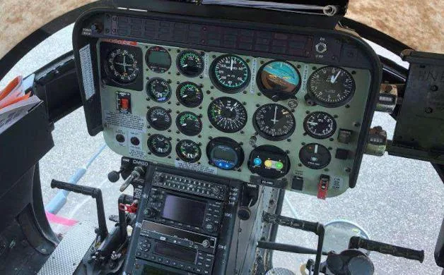 The cockpit of a helicopter showing the instrument panel with analog gauges and a digital display, and flight controls.