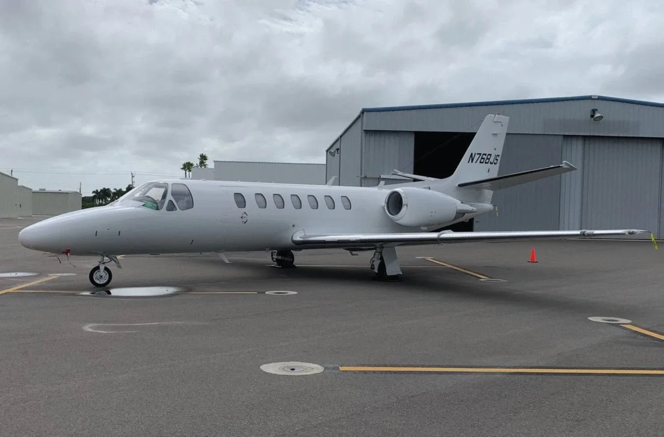 A white Cessna Citation business jet (N768JS) is parked on an airport tarmac next to a hangar under a cloudy sky.