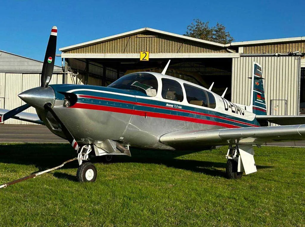 A Mooney M20J aircraft with a silver, blue, and red paint scheme parked on grass in front of a hangar.