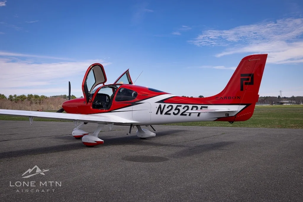 A rear-side view of a red and white Cirrus SR20 aircraft with its doors open, parked on an airfield.