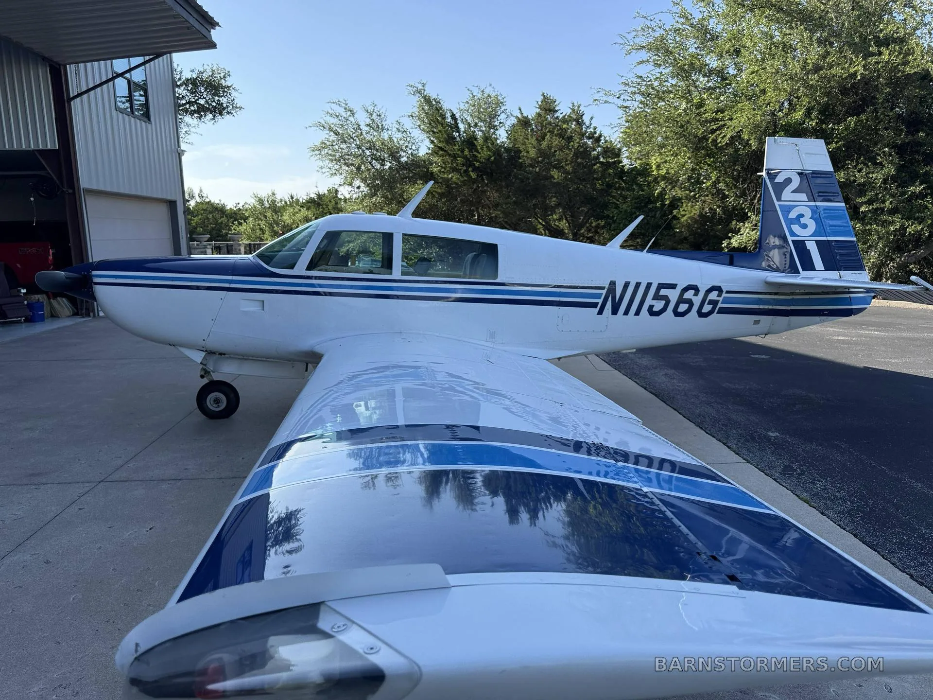 A white and blue single-engine aircraft (N156G) with blue stripes on the wing, parked next to a hangar.
