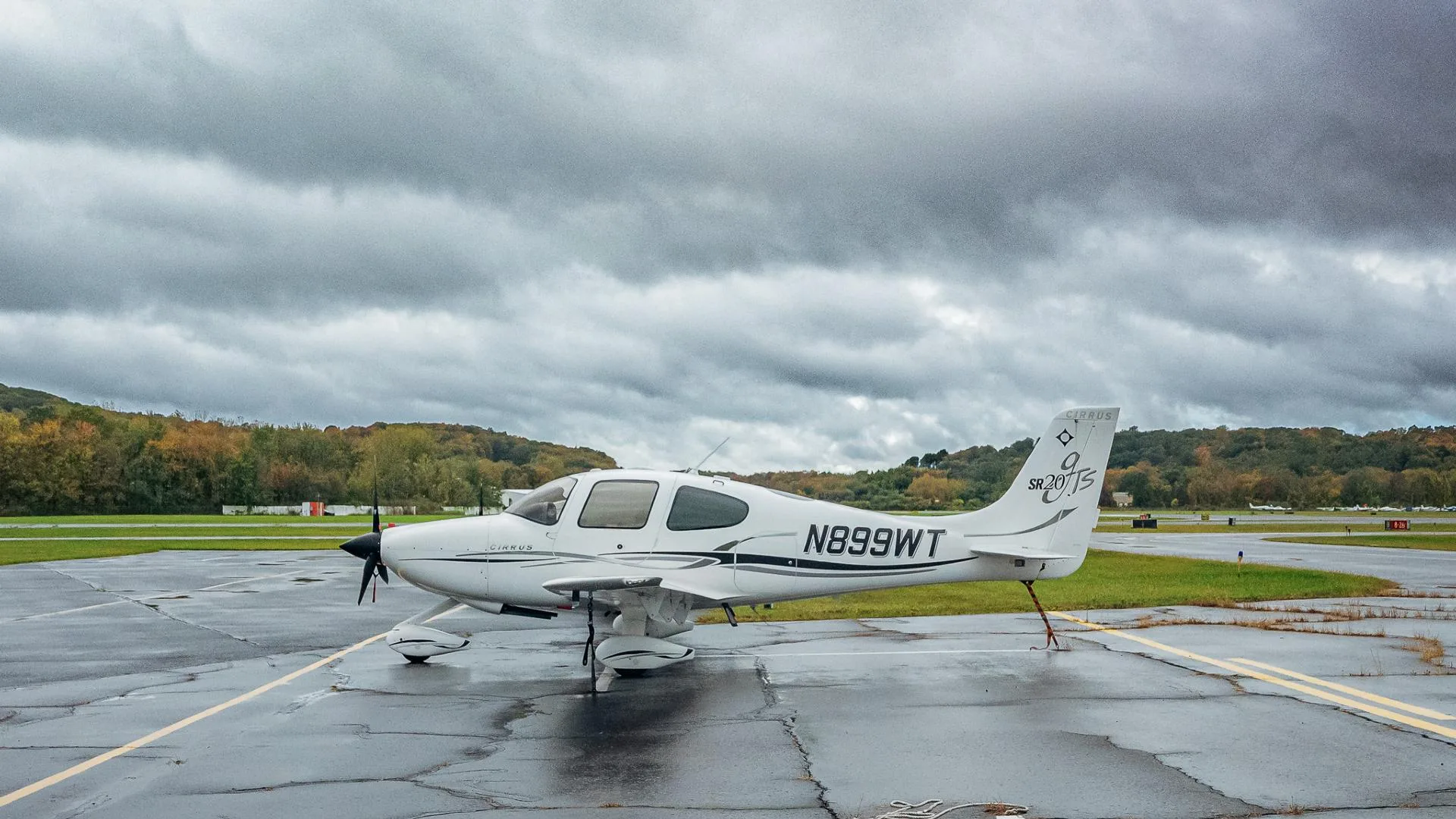 A white Cirrus SR20 aircraft with black and grey accents, registration N899WT, parked on a wet tarmac under a cloudy sky.