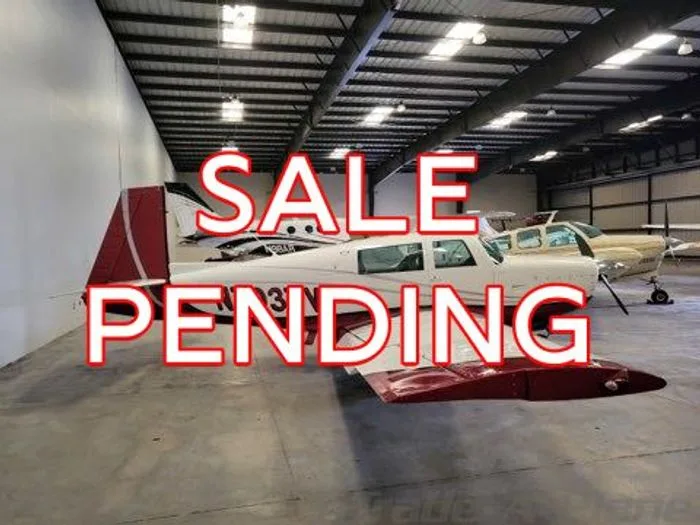 A white single-engine aircraft with red tail and wingtips is parked in a hangar, with another aircraft in the background.