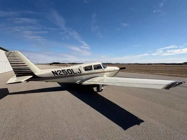 A cream-colored Piper Comanche 250 aircraft with brown stripes, seen from the rear-left, parked on an airfield under a blue sky.