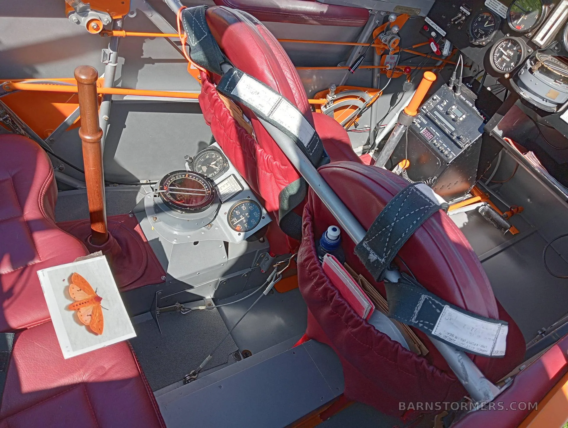 The interior of a biplane shows two red leather seats with harnesses, a compass, and a control stick.