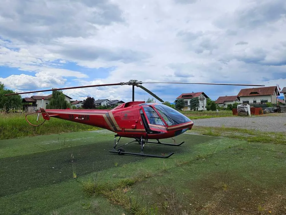 A red helicopter with gold stripes and an open door is parked on a green helipad, with houses and trees in the background.