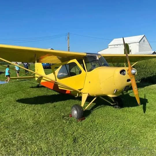 A yellow and orange single-engine high-wing aircraft is parked on a grassy field under a clear blue sky.