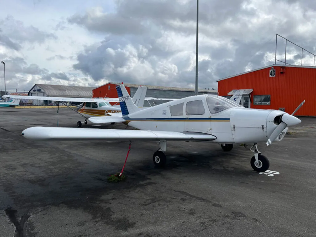 A white single-engine propeller aircraft with blue and gold stripes, tail number C-GCEP, parked on an airport tarmac with hangars in the background.