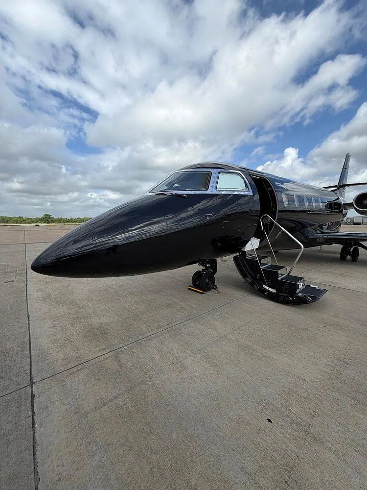 A sleek black business jet with its main door open and stairs extended, parked on an airfield under a partly cloudy sky.