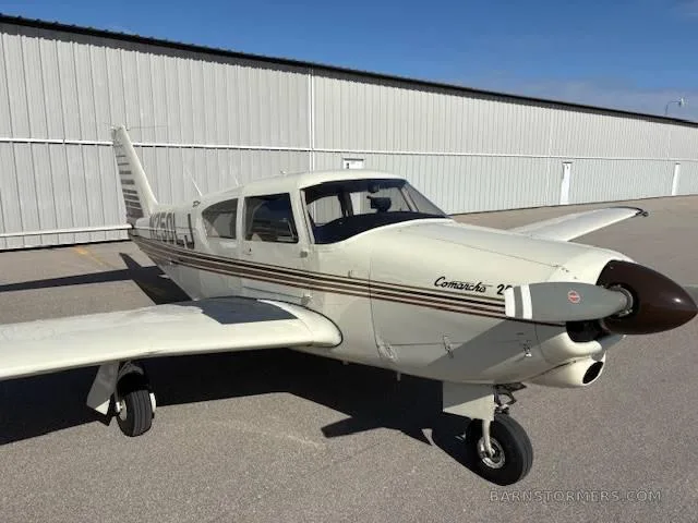 A cream-colored Piper Comanche 250 aircraft with brown stripes, parked on an asphalt apron in front of a hangar.