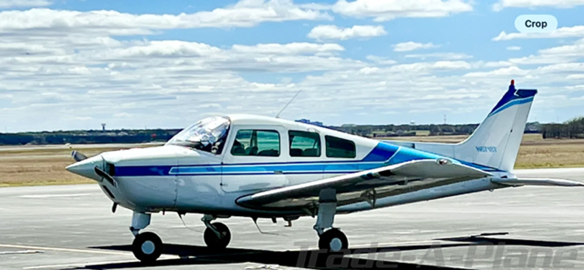 A white Beechcraft Sundowner N6718T with blue stripes on a tarmac under a cloudy sky, viewed from the side.