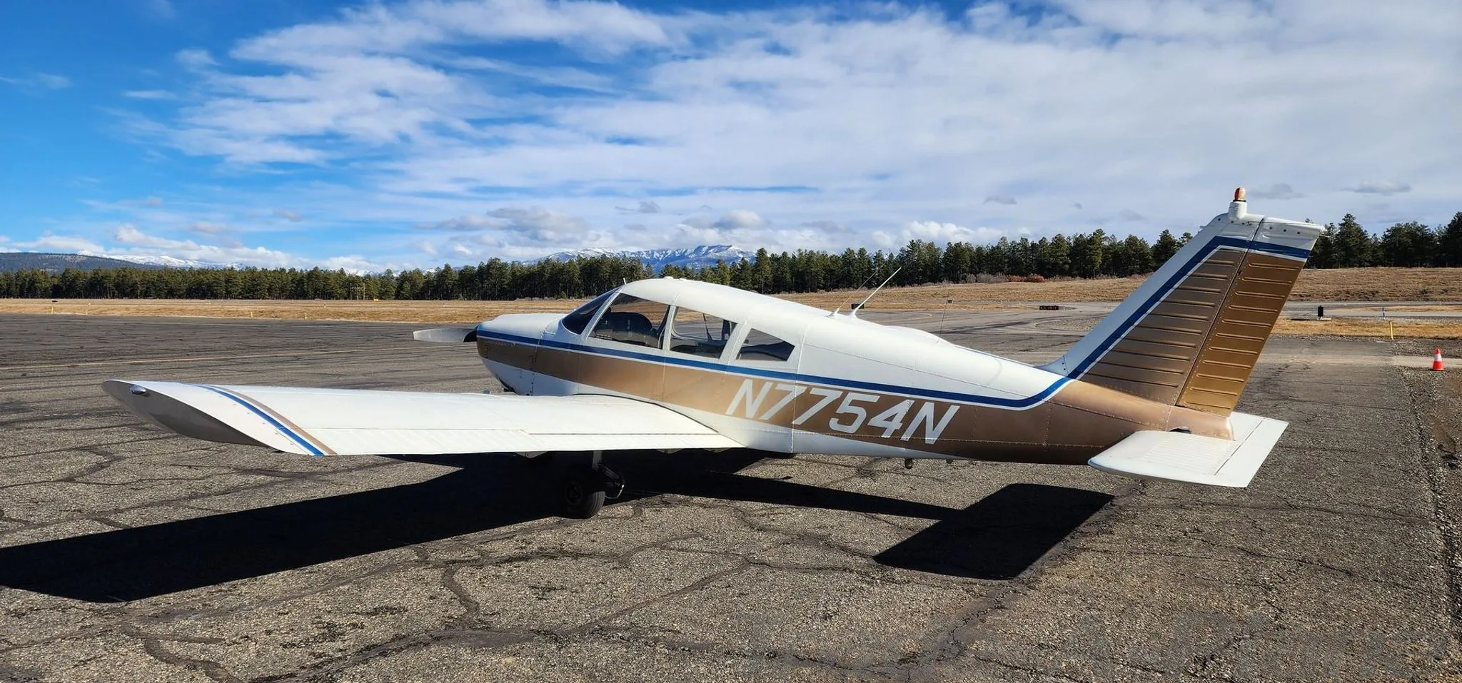 A white and gold Piper Cherokee 140 (N7754N) parked on an asphalt ramp with mountains in the background.