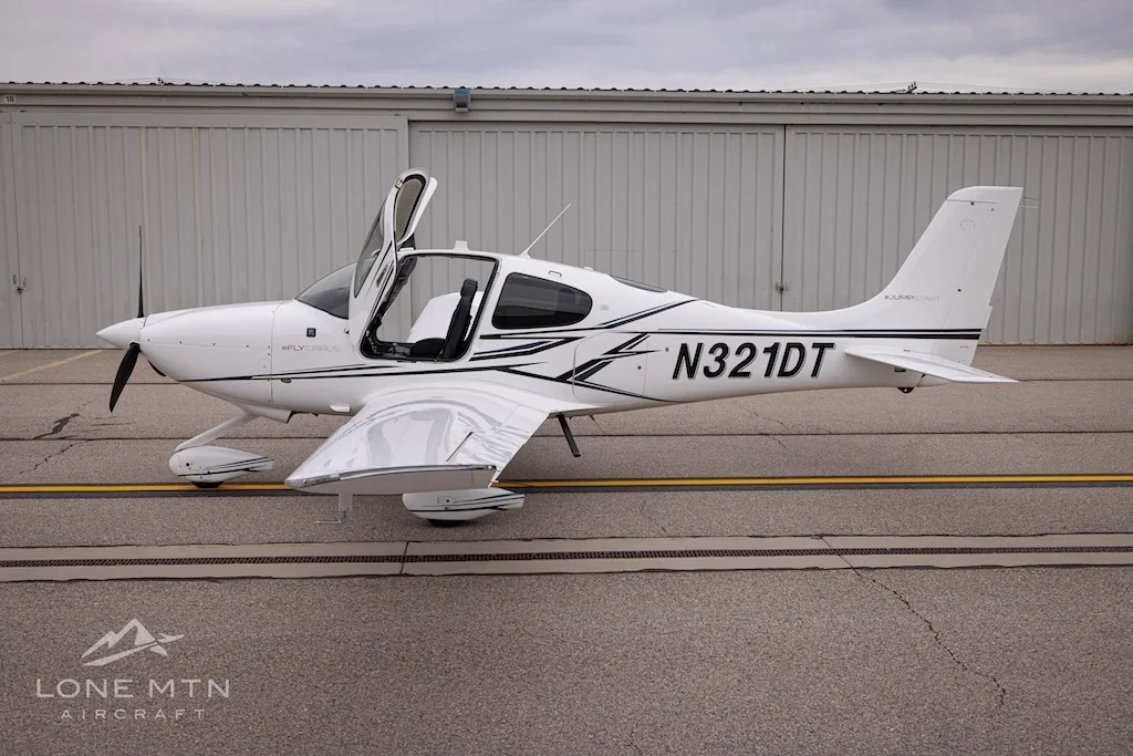 Side view of a white Cirrus SR22 aircraft with black and grey stripes, doors open, parked on an asphalt ramp.