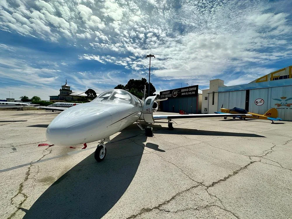 A white Cessna CitationJet aircraft with a red stripe is parked on an airport tarmac, viewed from the front-left, under a cloudy sky.
