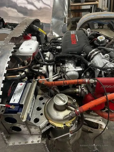Detailed view of an aircraft engine bay, showing the Rotax engine, battery, and various hoses and components.