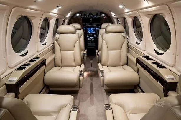 The aircraft cabin features light beige leather seats arranged in a club configuration, with windows along the sides and a view towards the cockpit.