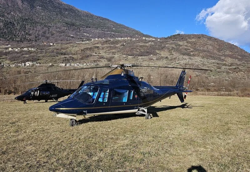 Two AgustaWestland AW109SP helicopters are parked on a grassy field with a mountain in the background.