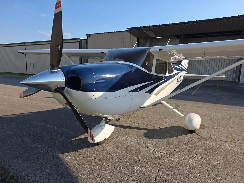 Front-quarter view of a white and blue Cessna 182 aircraft with registration N147AJ parked on an asphalt ramp.