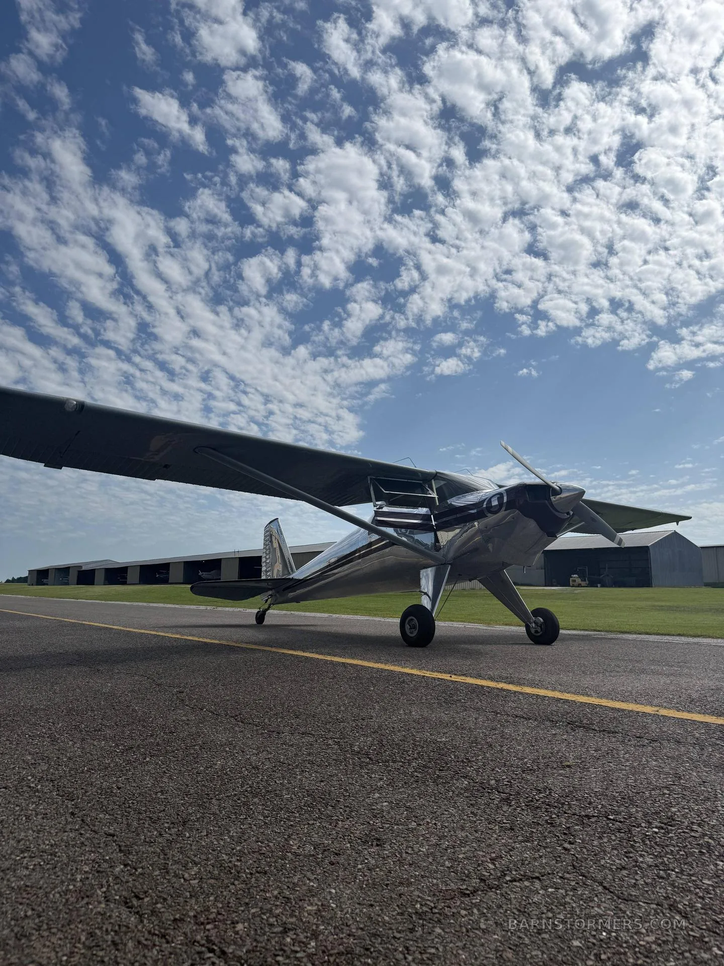A shiny silver high-wing aircraft with a dark nose and propeller, parked on an asphalt runway under a blue sky with scattered clouds.
