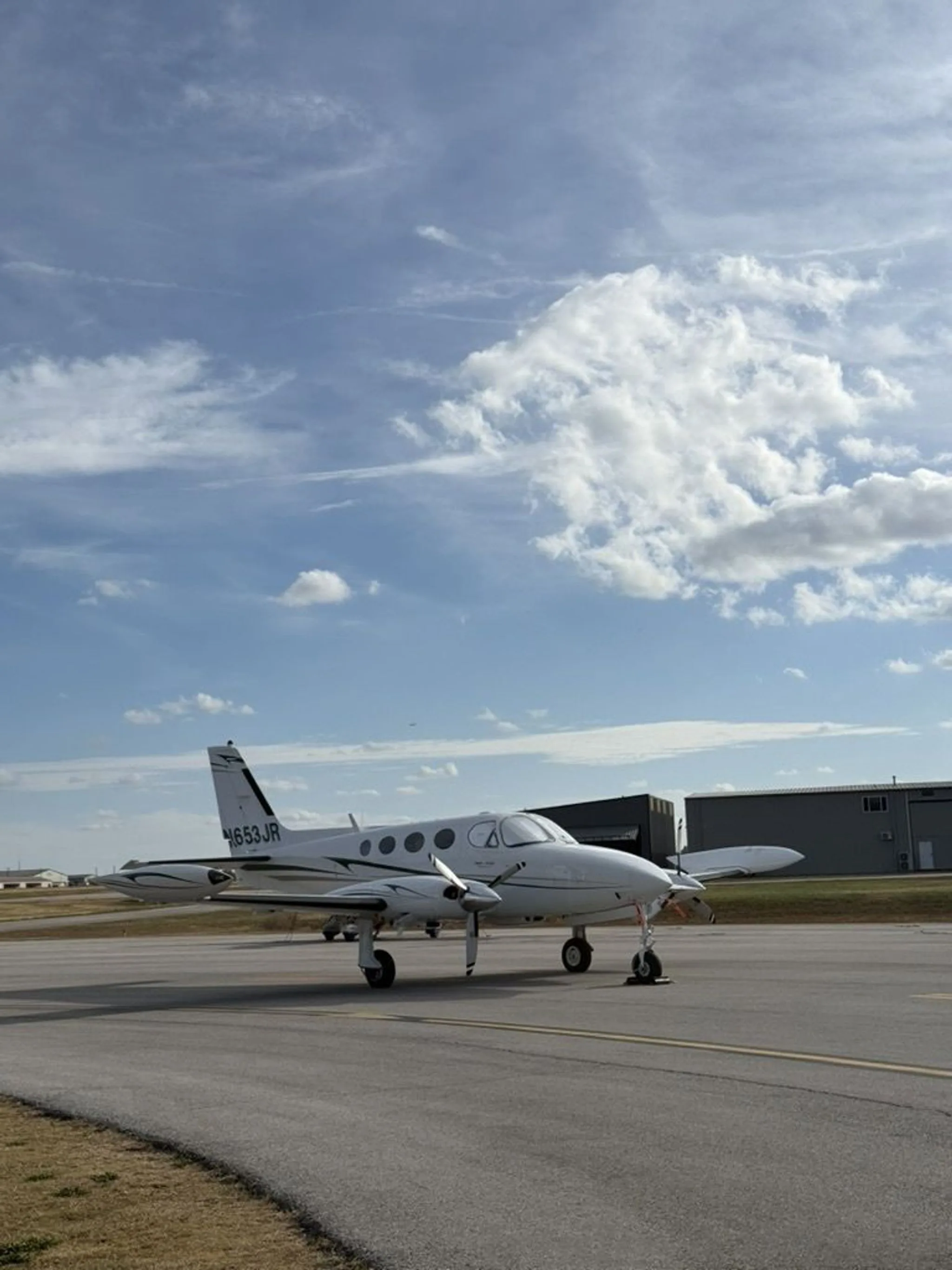A white twin-engine aircraft, N653JR, is parked on an airport tarmac under a partly cloudy blue sky.