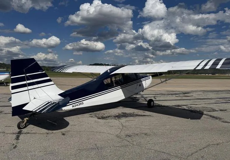 A white and dark blue high-wing aircraft, registration N343RS, parked on a tarmac from a rear-quarter view under a partly cloudy sky.
