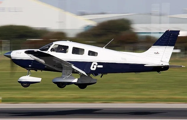 A white and dark blue Piper Archer II aircraft with registration G- is shown in flight over a runway.