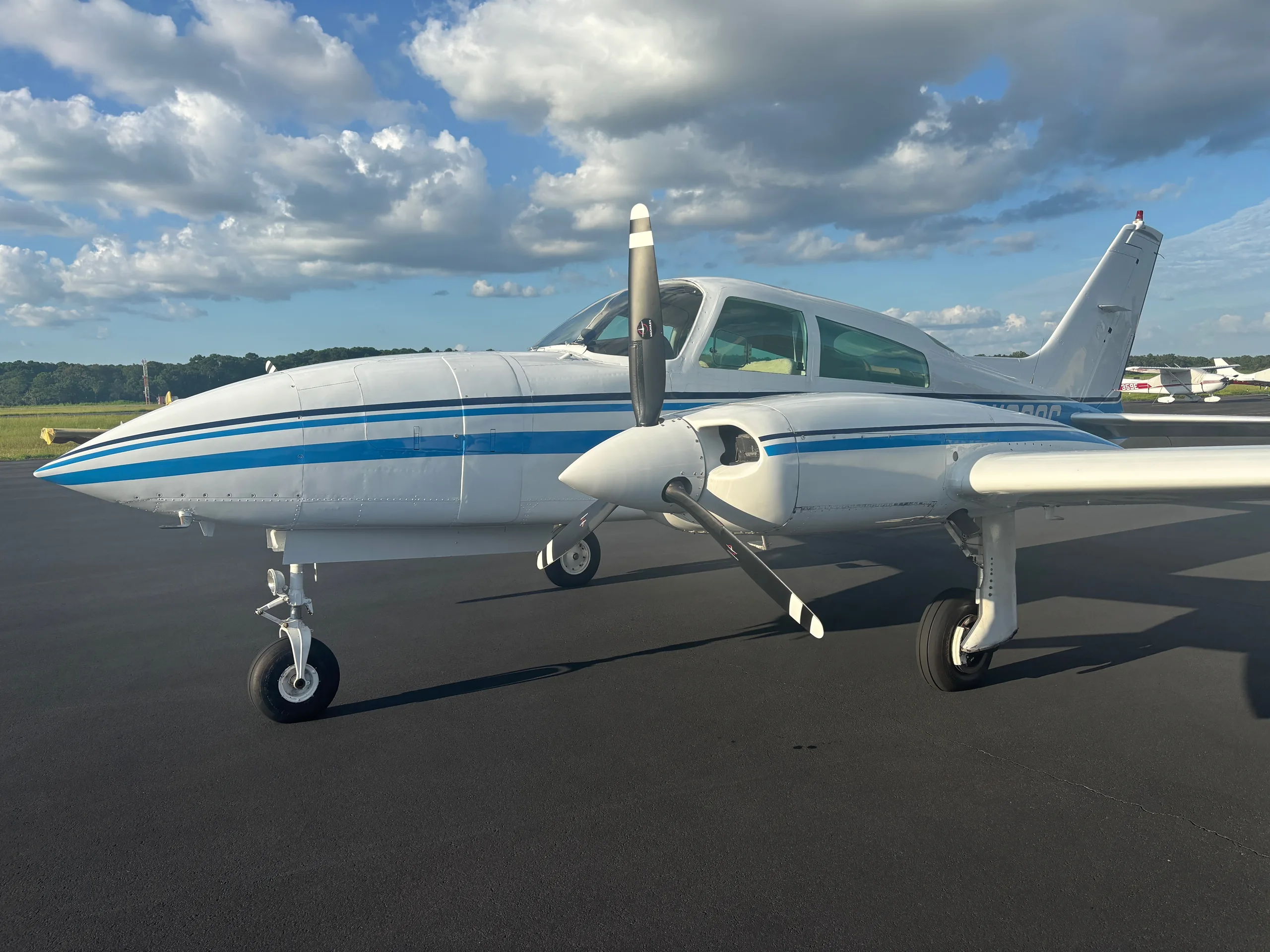 A white twin-engine aircraft with blue stripes parked on an asphalt runway, viewed from the front-right.