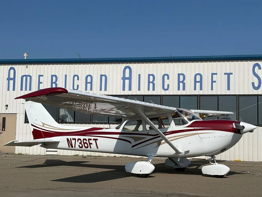 A white and maroon Cessna 172 with gold accents, parked in front of a building labeled "American Aircraft Sales".