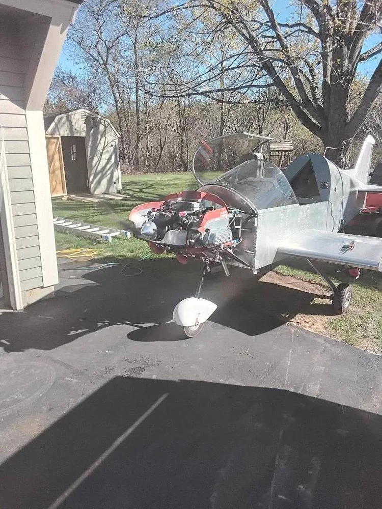 A partially assembled light aircraft, unpainted and with its engine exposed, parked on a driveway in a residential yard.