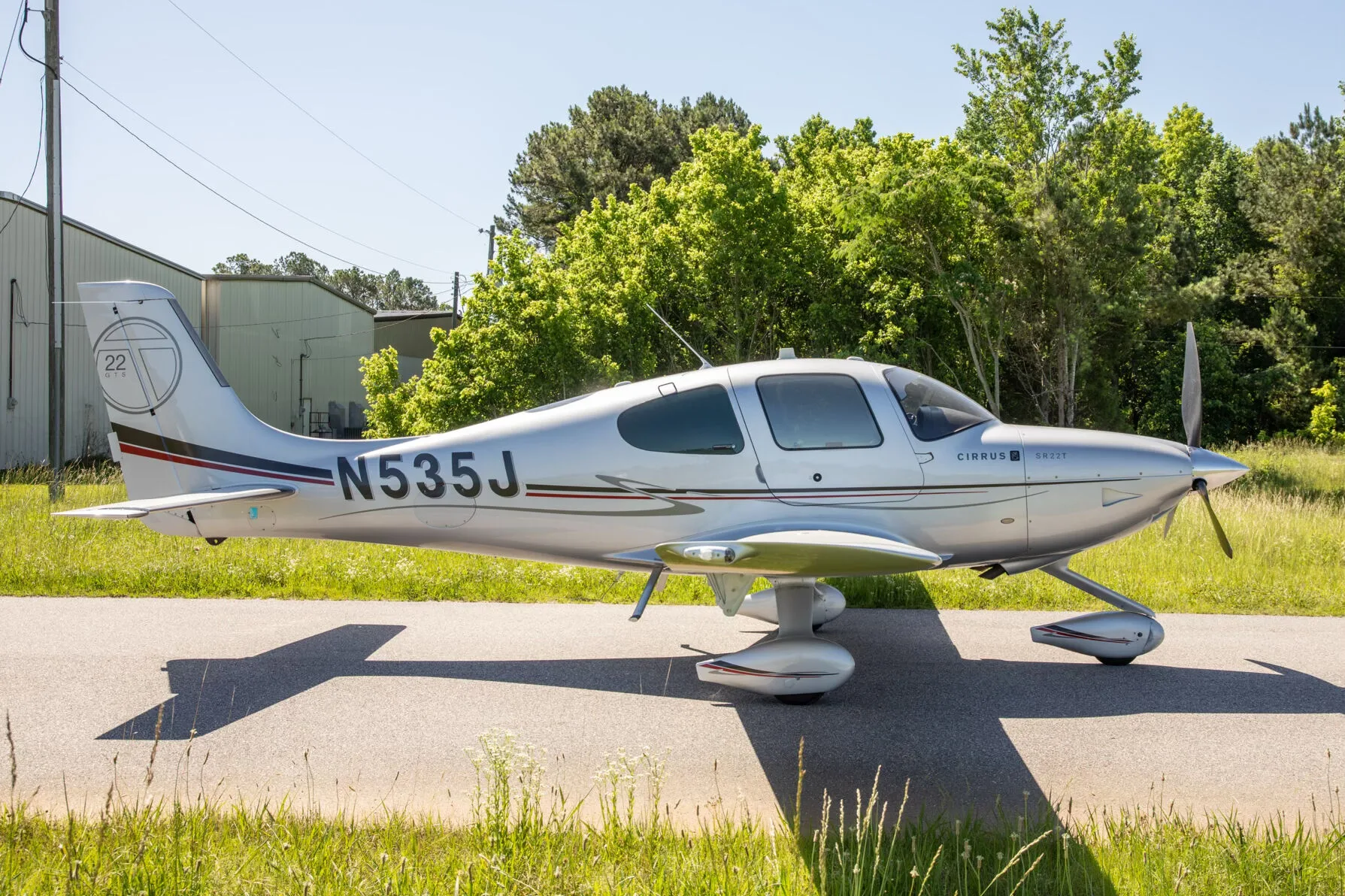 A silver Cirrus SR22T aircraft with registration N535J is parked on an asphalt strip with hangars and trees in the background.