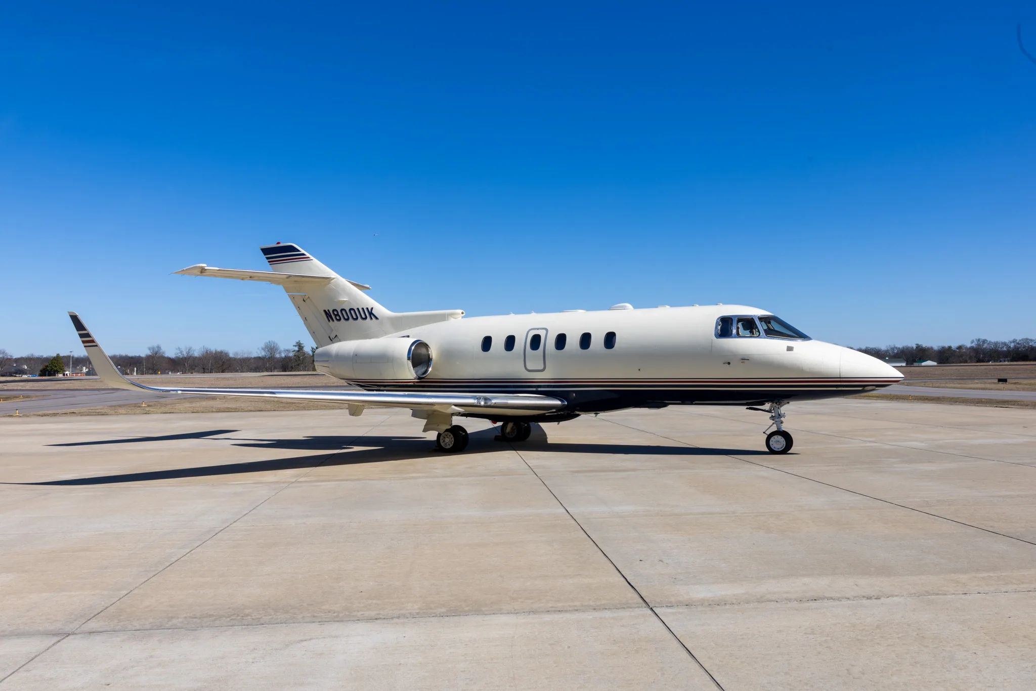 A white and blue business jet, N800UK, is parked on a concrete tarmac under a clear blue sky.