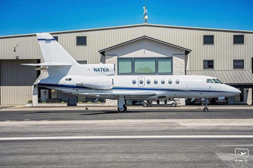 A white Dassault Falcon 900EX business jet with blue stripes on the fuselage and tail, parked in front of a hangar.
