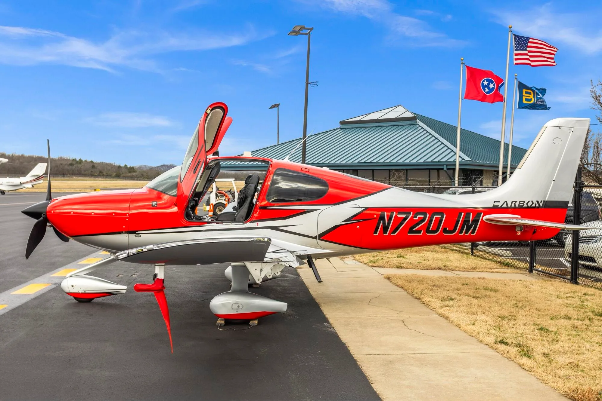 A side view of a red and silver Cirrus SR22 aircraft with its gull-wing doors open, parked on an airport tarmac.