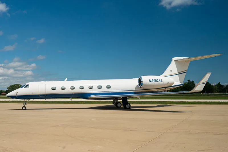 A white Gulfstream G550 business jet with blue and gold stripes on the tarmac under a blue sky.