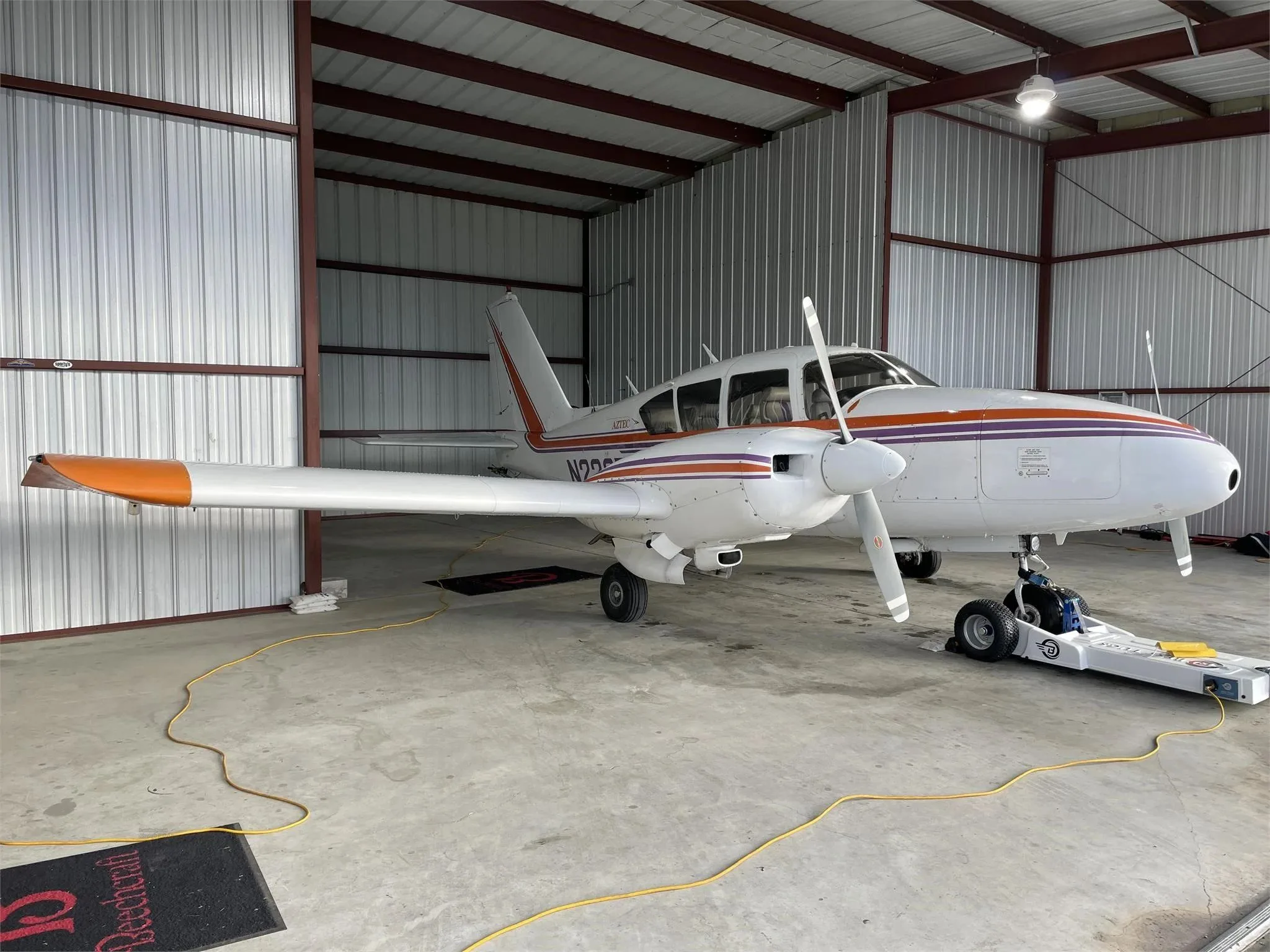 A white Piper Aztec twin-engine aircraft with orange and purple stripes parked in a hangar with a tow bar attached to the nose wheel.