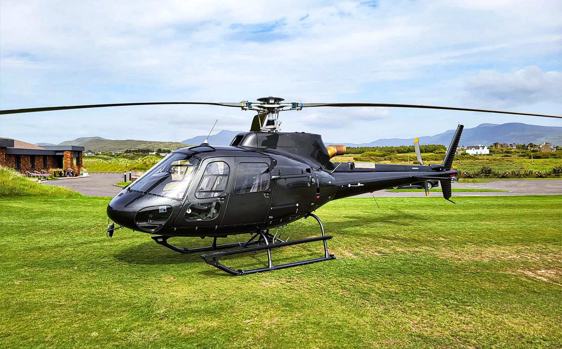 A sleek black helicopter, likely an Airbus H125, is parked on a vibrant green lawn with a modern building and distant mountains under a cloudy sky.