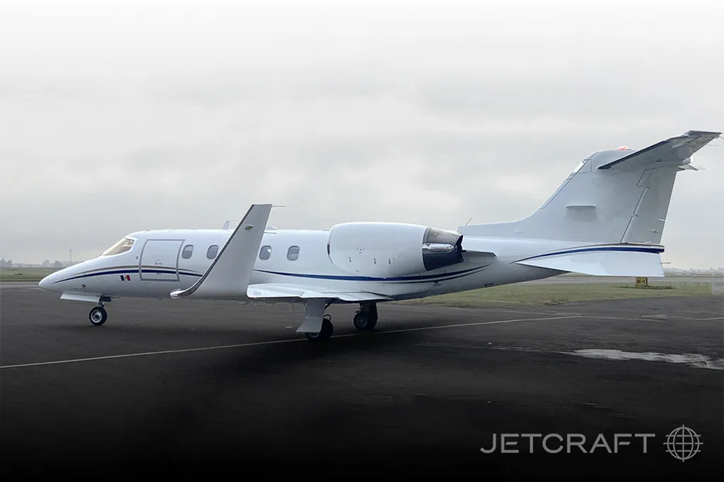 A white business jet, likely a Learjet, with blue accent stripes on the tail, parked on an asphalt tarmac under an overcast sky.