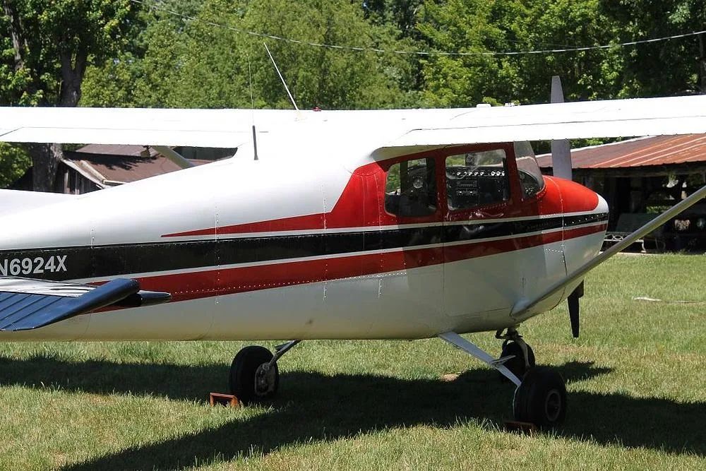 A mid-section side view of a red and white Cessna 172 (N6924X) parked on grass near trees.