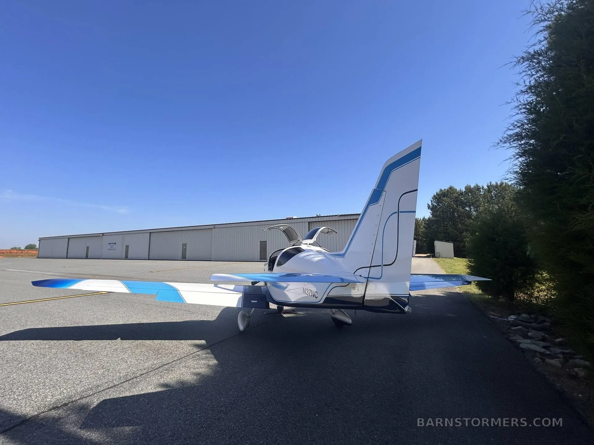 A white and blue single-engine aircraft with registration N37NC is parked on an asphalt tarmac with its canopy open, viewed from the rear.