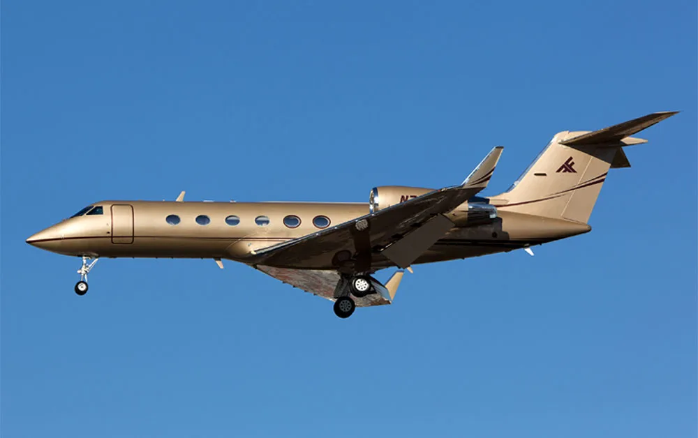 A gold Gulfstream business jet with landing gear extended flies against a clear blue sky.